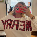White crewneck sweatshirt with red plaid appliqué "MERRY" lettering, styled with red ribbed neckline trim.
