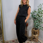 Black sleeveless muscle-tee jumpsuit with drawstring waist, styled with silver sneakers and gold bracelet.