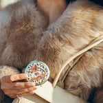 Teddy bear compact mirror held by woman wearing fur coat, paired with cream Chanel handbag.