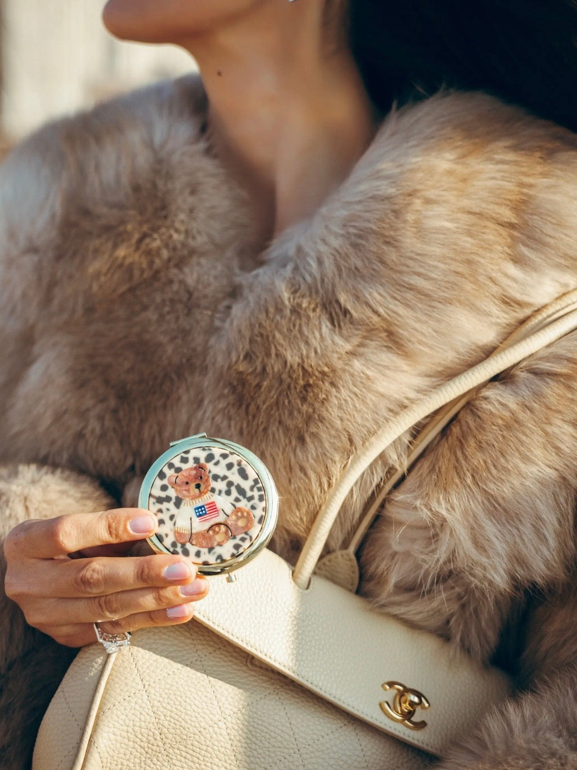 Teddy bear compact mirror held by woman wearing fur coat, paired with cream Chanel handbag.