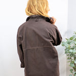 Back view of an oversized dark brown utility jacket with contrast stitching, worn with blue jeans.