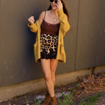Woman wearing a leopard print mini skirt with black lace hem, brown cami, and mustard cardigan