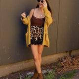 Woman wearing a leopard print mini skirt with black lace hem, brown cami, and mustard cardigan