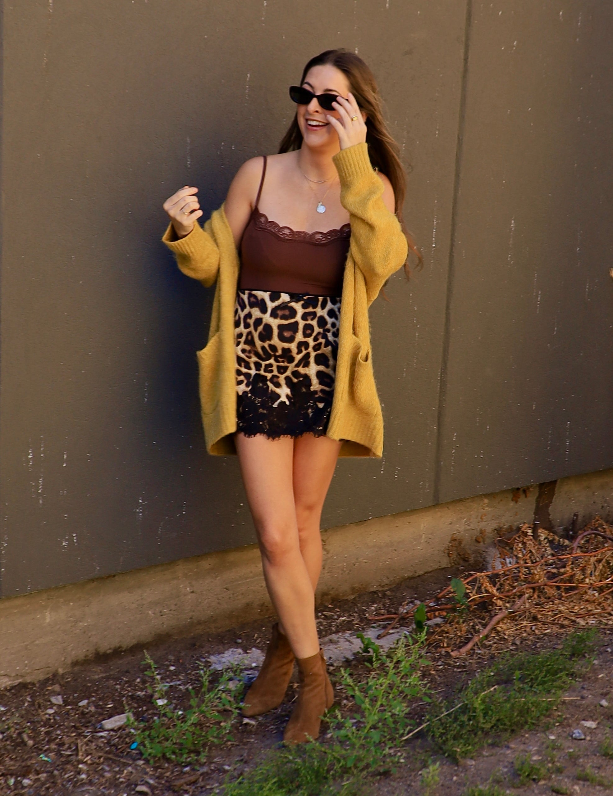 Woman wearing a leopard print mini skirt with black lace hem, brown cami, and mustard cardigan