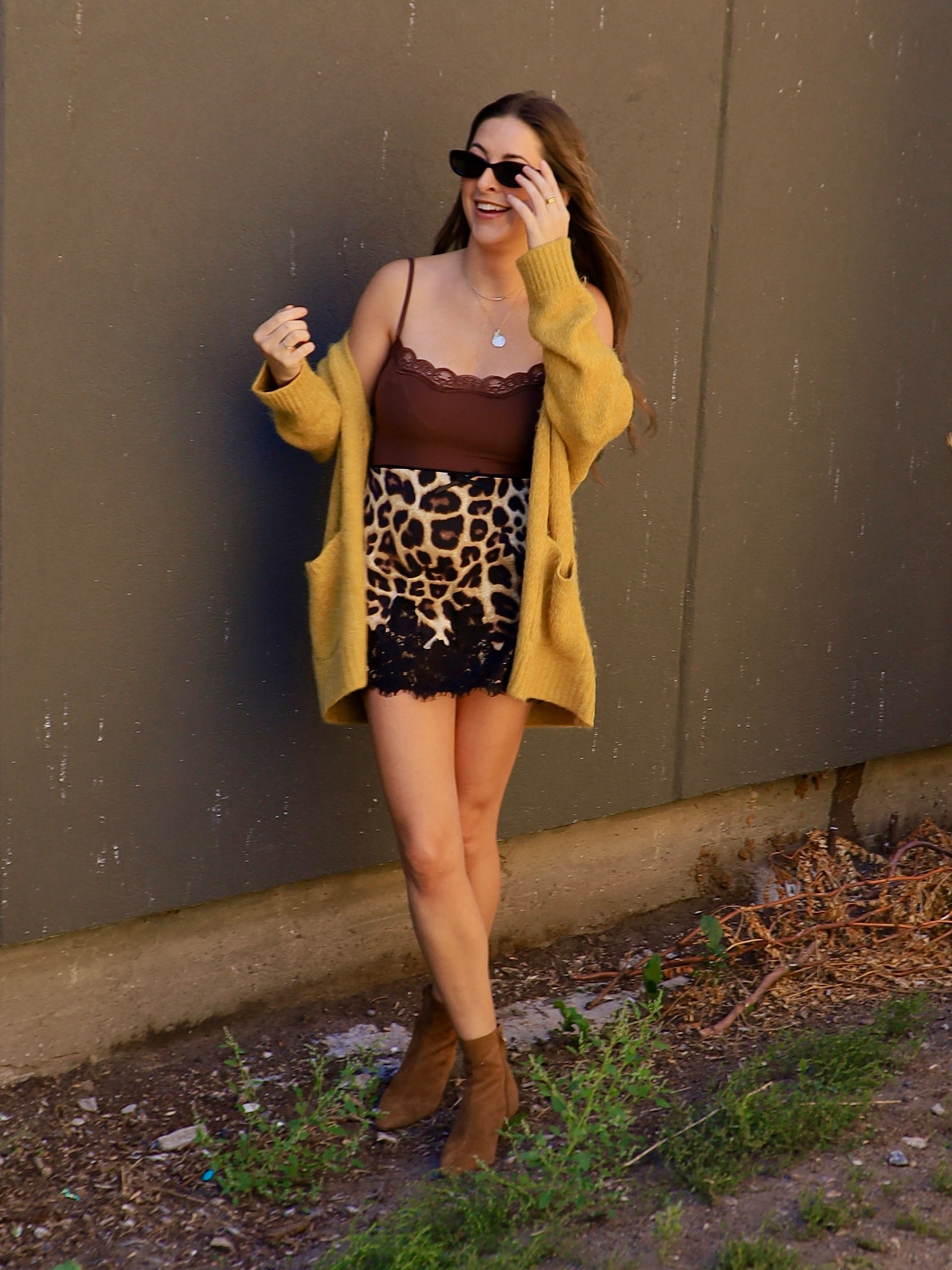 Woman wearing a leopard print mini skirt with black lace hem, brown cami, and mustard cardigan