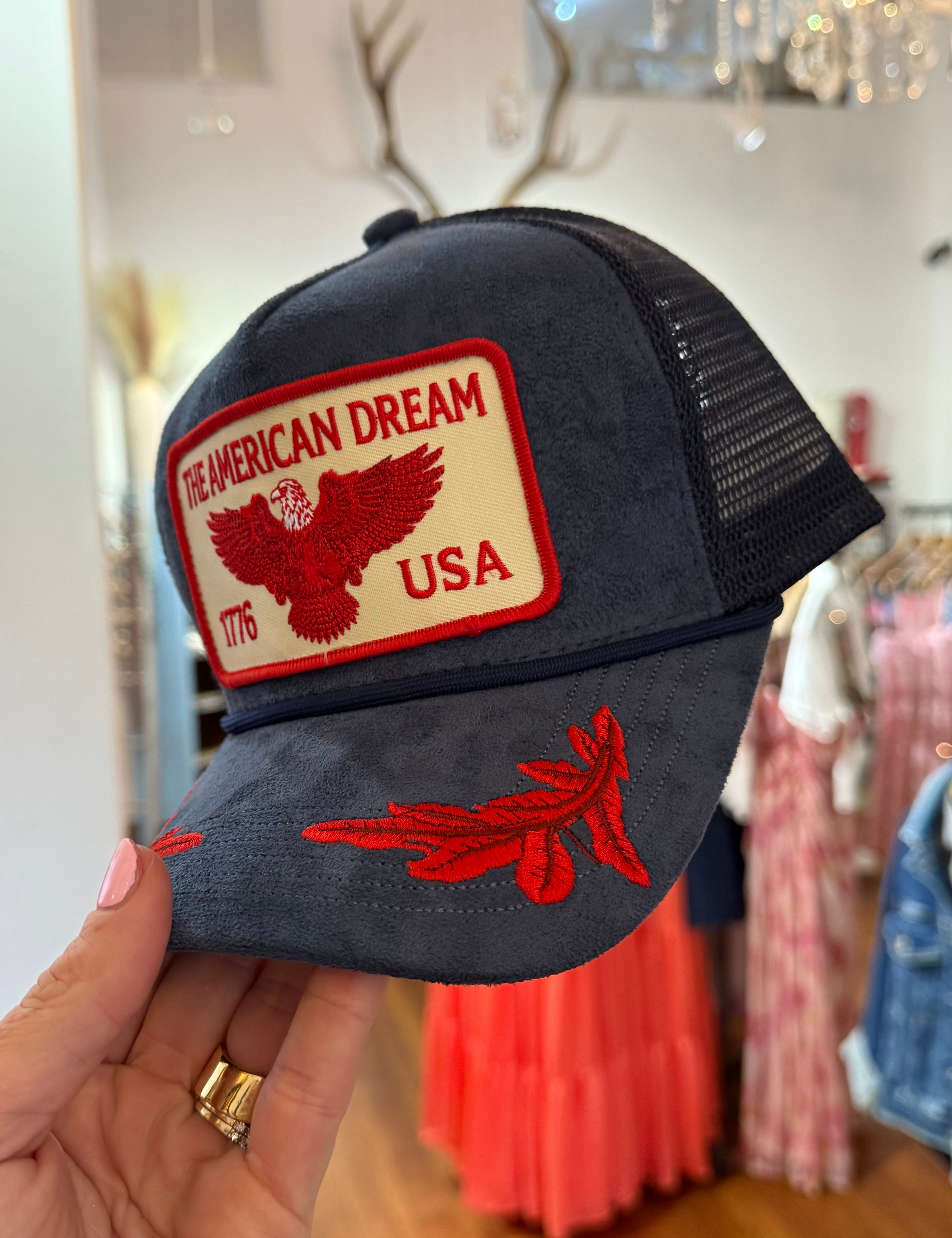 Navy blue suede trucker hat with red embroidered "The American Dream USA 1776" eagle patch and feather brim detail.