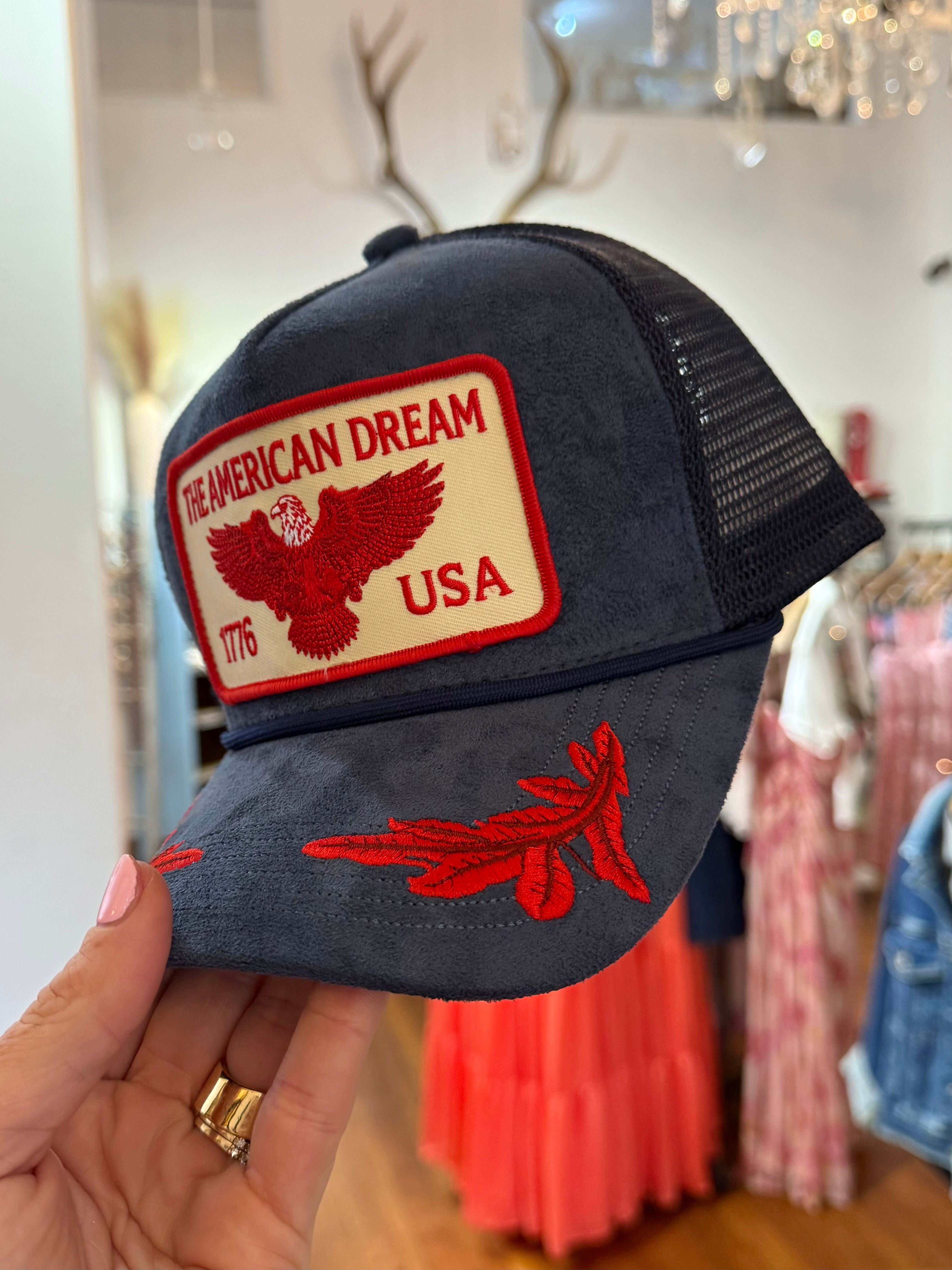 Navy blue suede trucker hat with red embroidered "The American Dream USA 1776" eagle patch and feather brim detail.