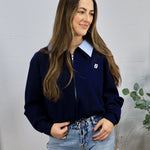 Navy zip-up cardigan with white logo, layered over blue striped collar shirt, paired with light wash jeans.