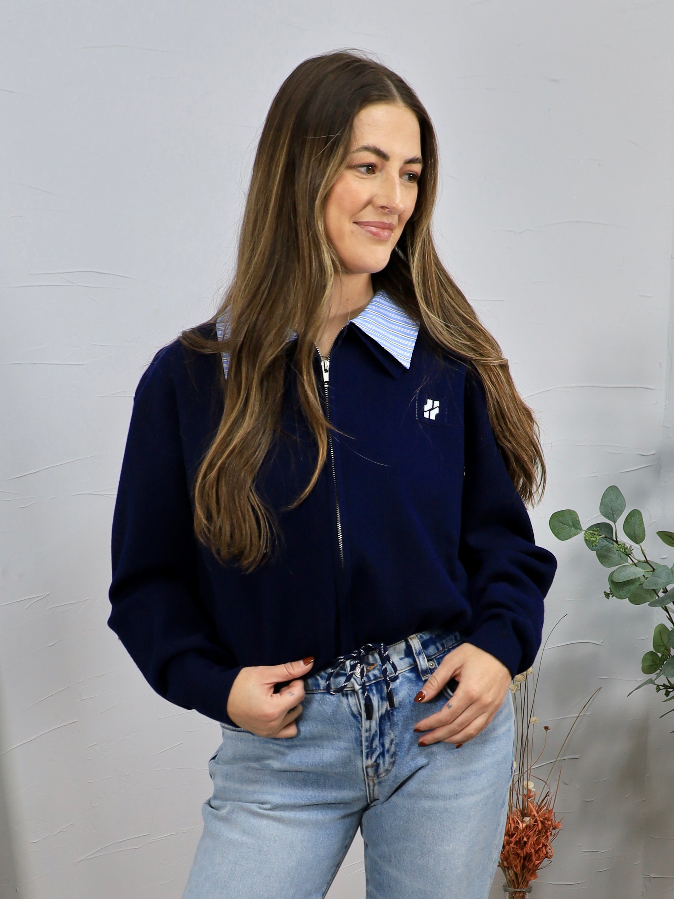 Navy zip-up cardigan with white logo, layered over blue striped collar shirt, paired with light wash jeans.