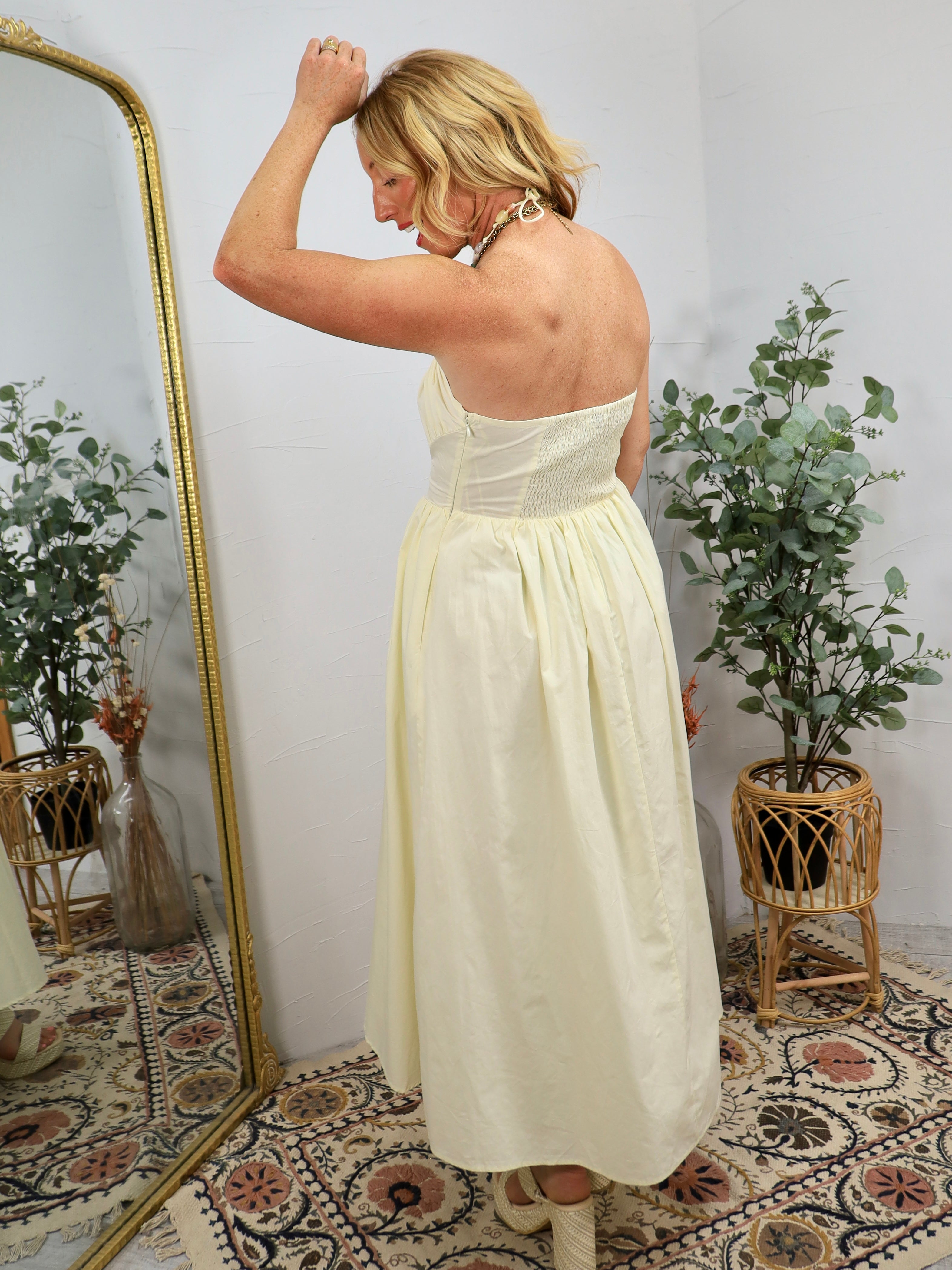 Woman in cream strapless maxi dress with smocked waist detail, styled with beaded earrings in bohemian room setting.
