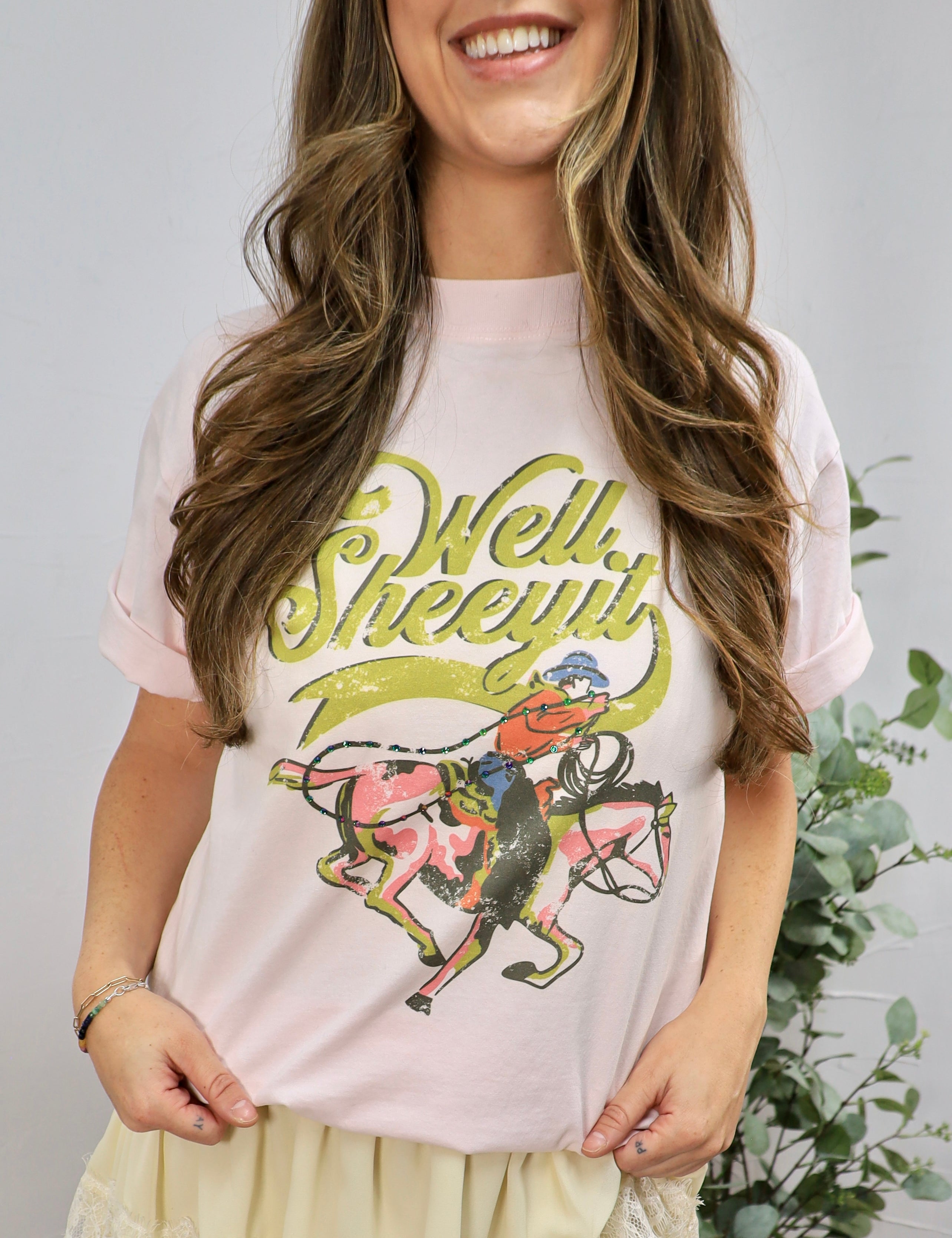 Woman wearing light pink graphic tee with vintage cowboy design and Well Sheeyit text