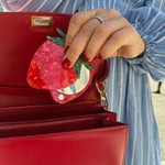 Strawberry-shaped red acrylic compact mirror being placed into an open red leather structured handbag.