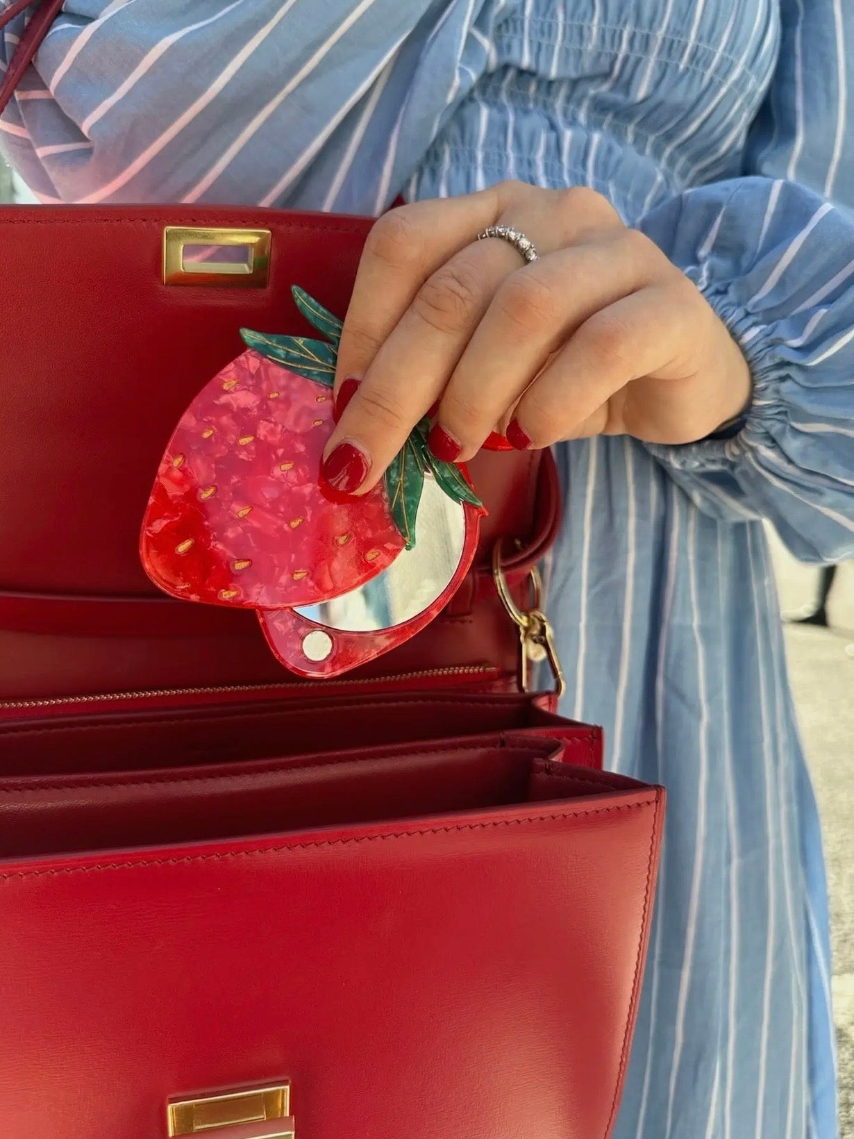 Strawberry-shaped red acrylic compact mirror being placed into an open red leather structured handbag.