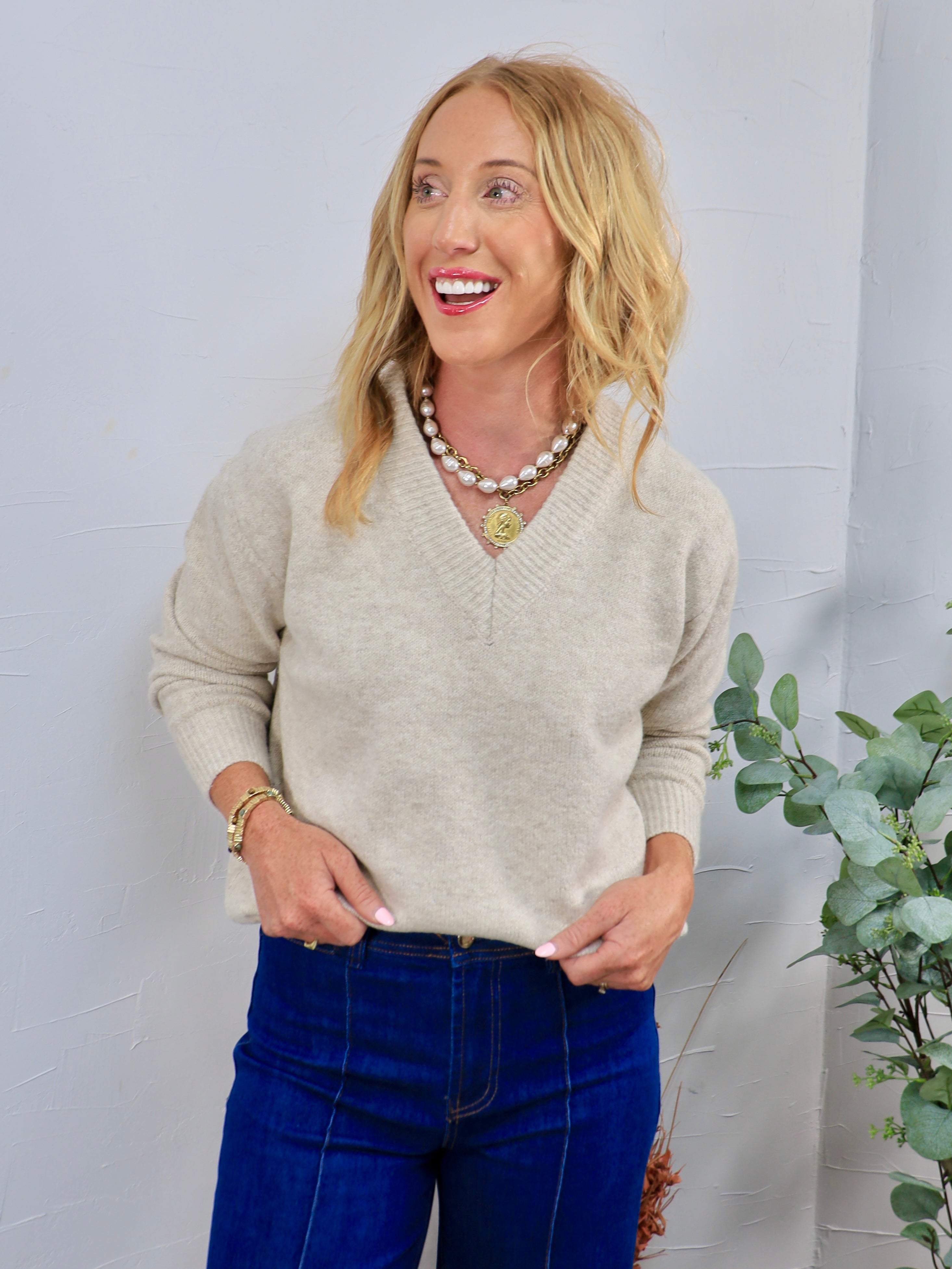 Beige v-neck knit sweater styled with dark blue jeans, gold coin necklace, and pearl chain layered necklace.