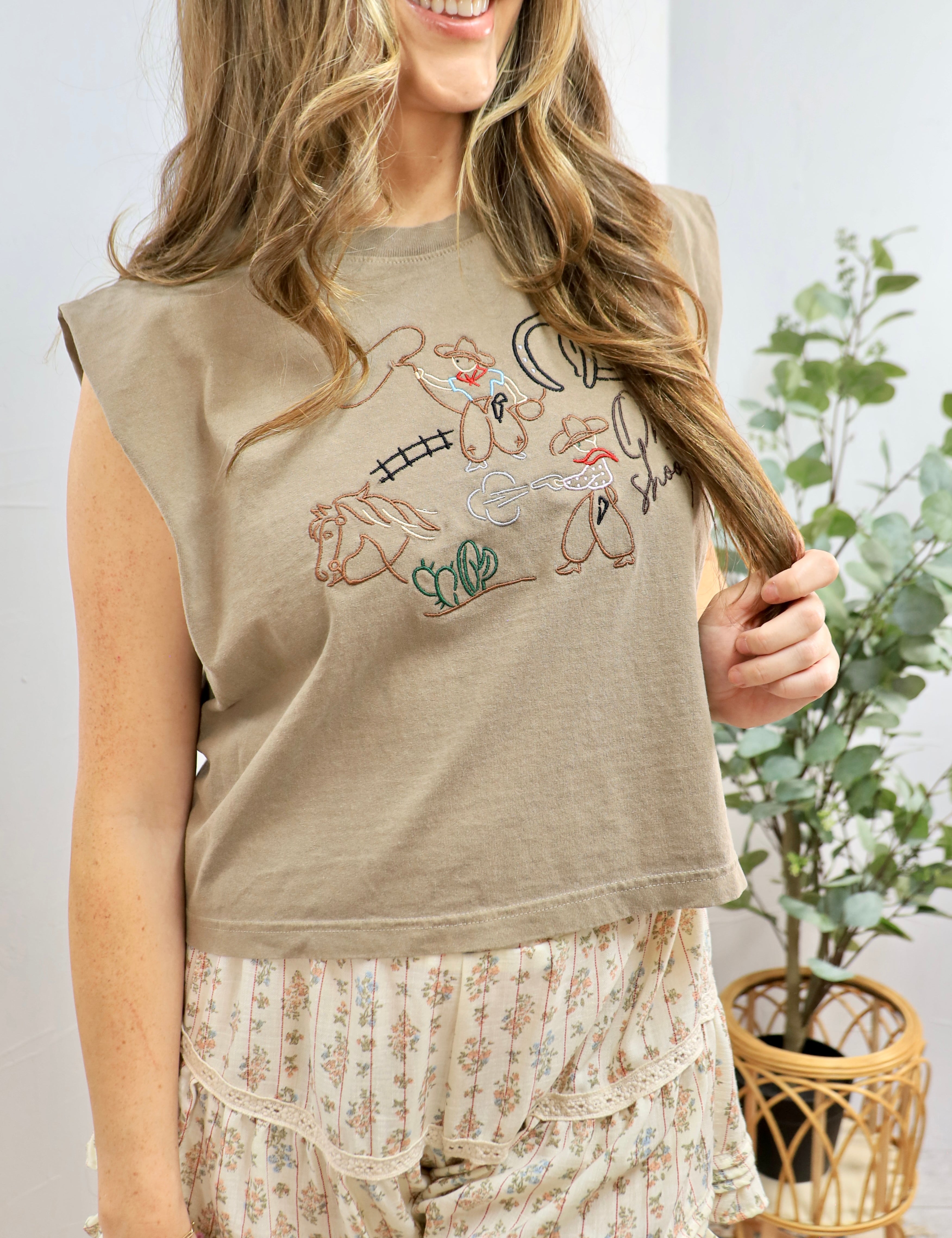 Woman wearing sage green sleeveless tee with colorful rodeo-themed embroidery, styled with floral midi skirt and blonde wavy 