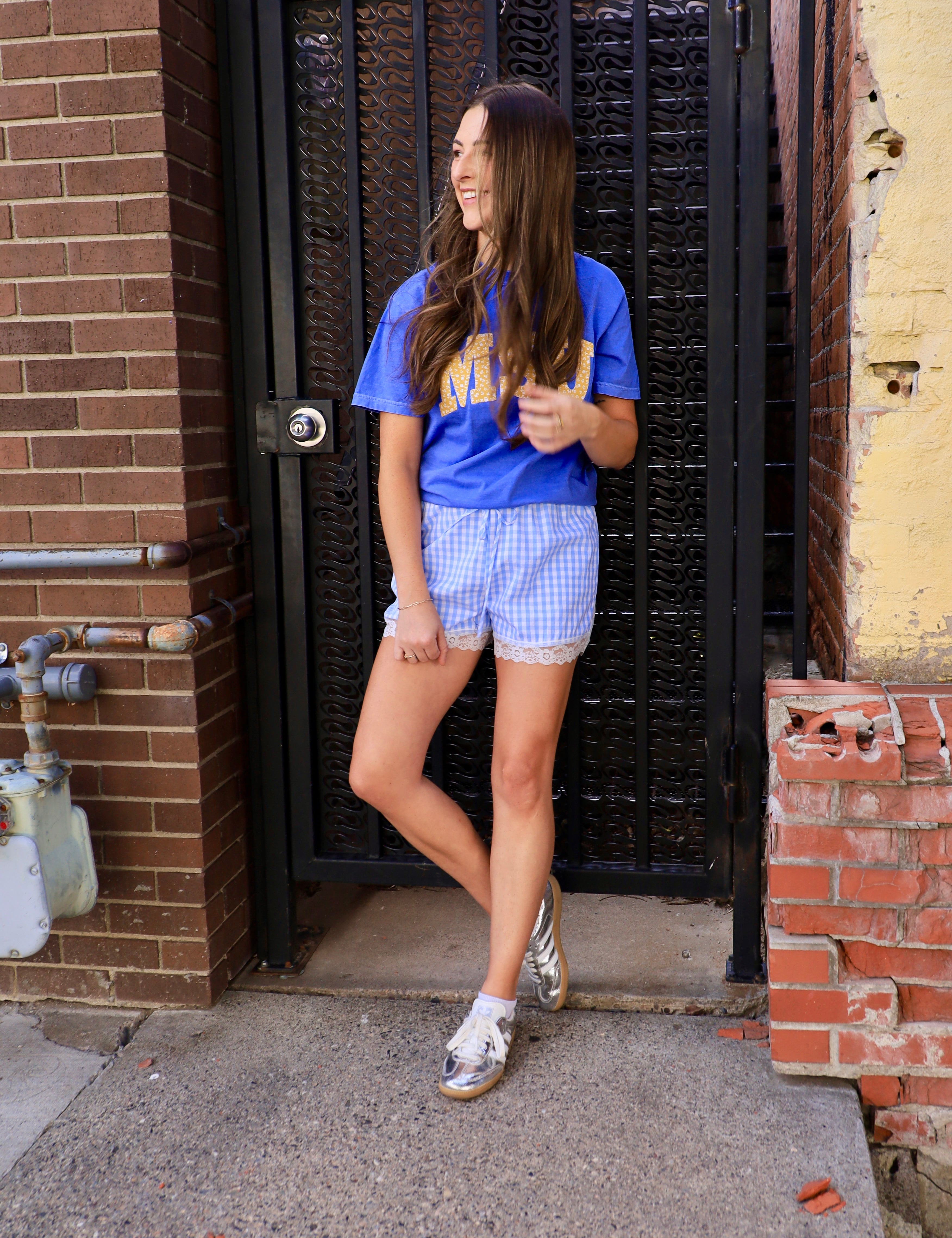 Woman wearing royal blue cropped MSU graphic tee with gold lettering paired with blue gingham shorts and silver sneakers