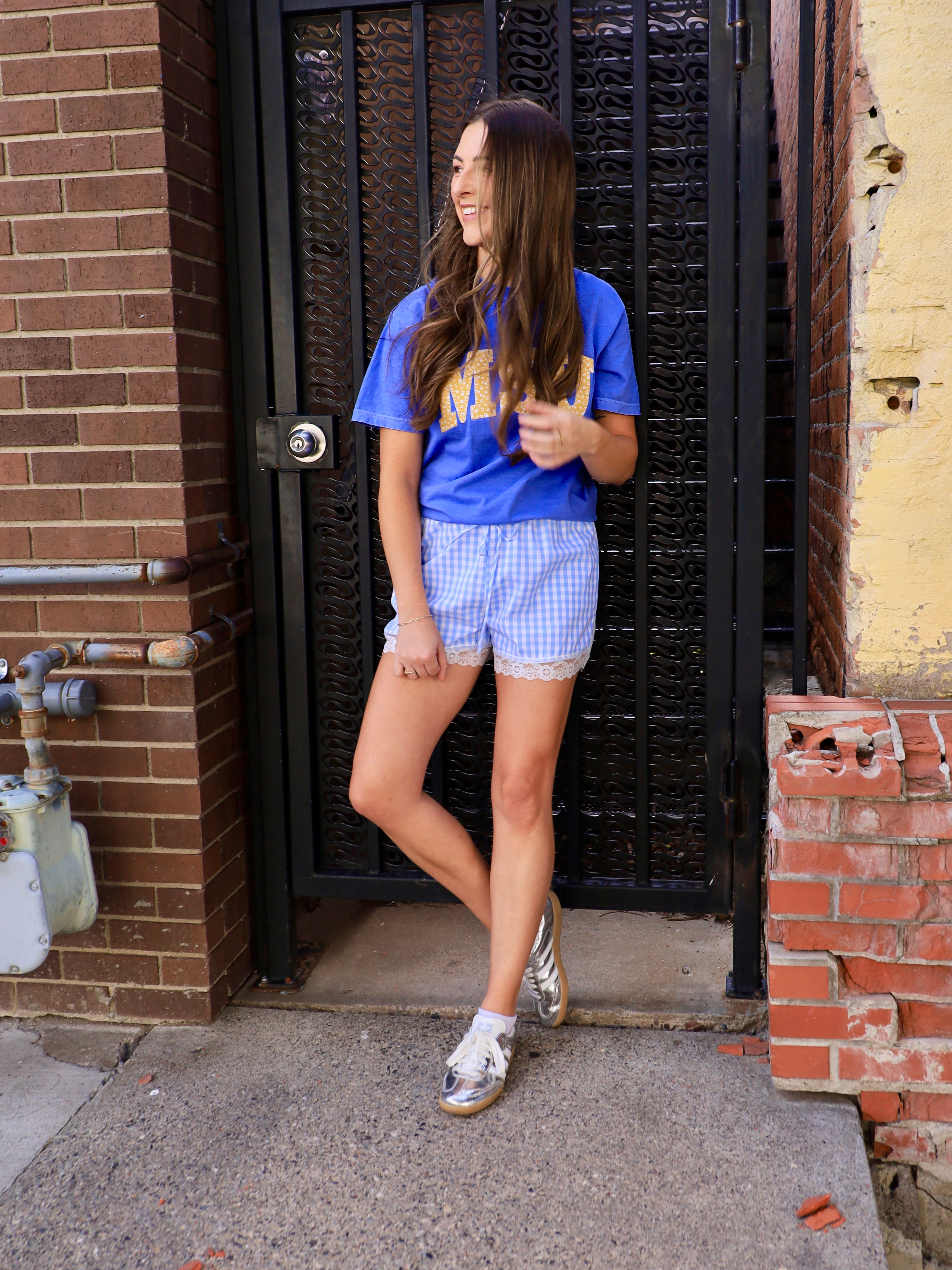 Woman wearing royal blue cropped MSU graphic tee with gold lettering paired with blue gingham shorts and silver sneakers