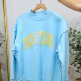 Light blue mockneck sweatshirt with "MONTANA" text in yellow and patterned fabric letters, displayed on wooden hanger.