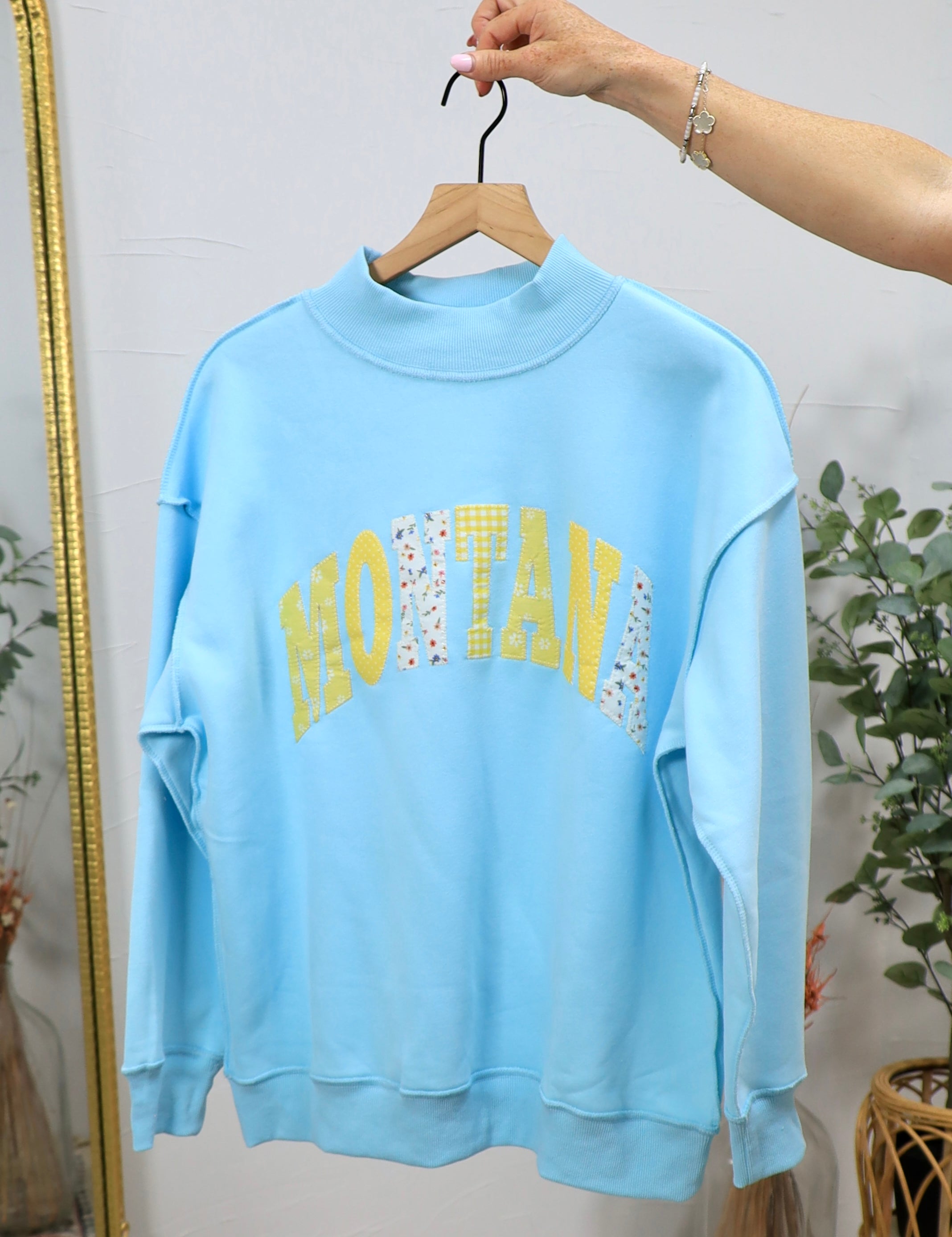 Light blue mockneck sweatshirt with "MONTANA" text in yellow and patterned fabric letters, displayed on wooden hanger.