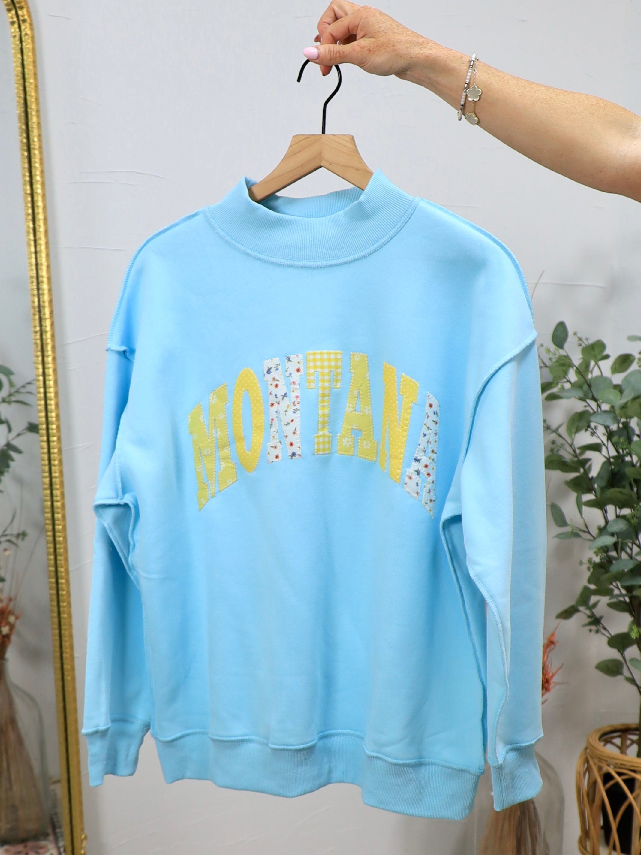 Light blue mockneck sweatshirt with "MONTANA" text in yellow and patterned fabric letters, displayed on wooden hanger.
