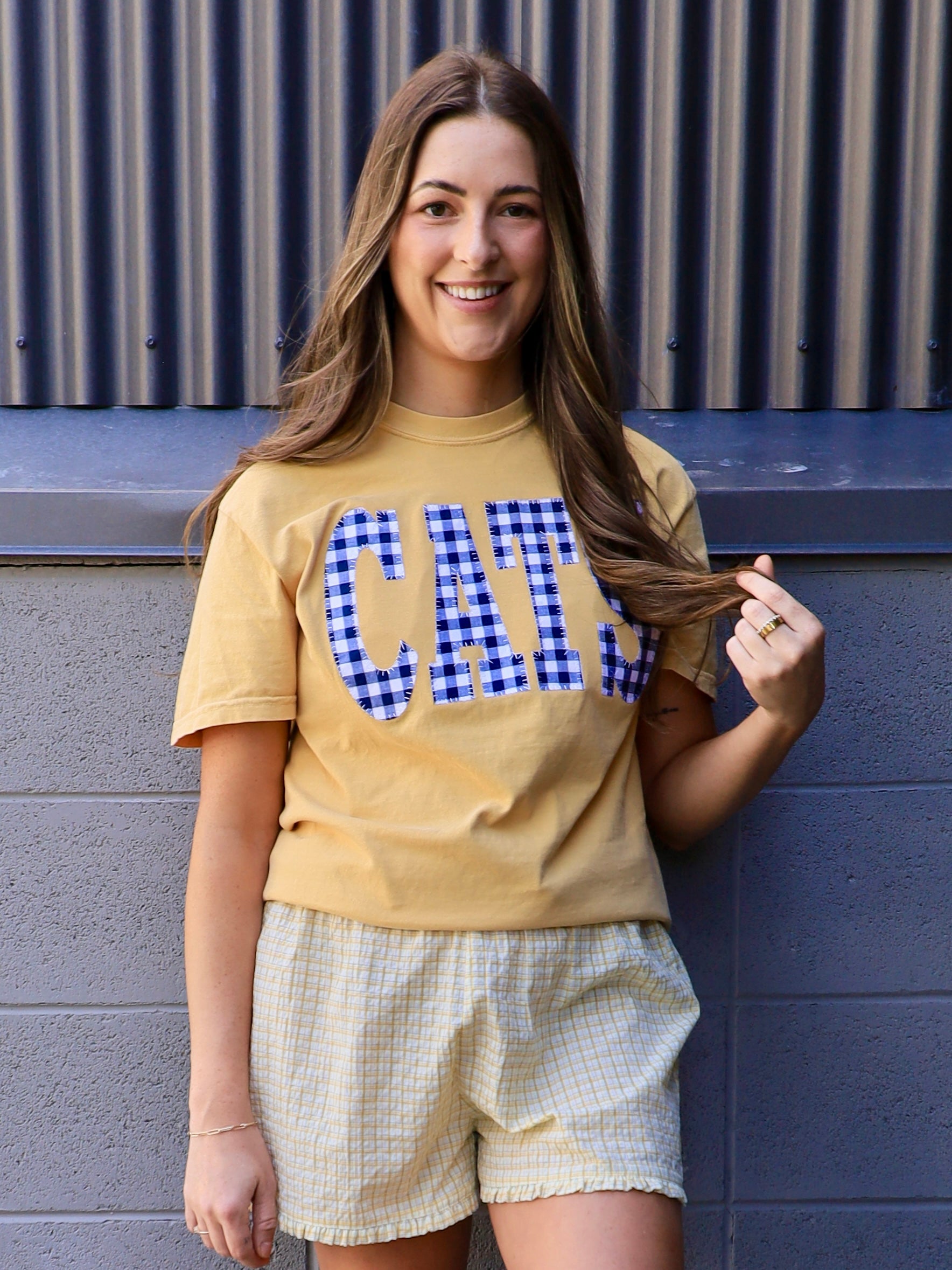 Yellow "CATS" gingham appliqué tee styled with matching plaid smocked shorts, worn by smiling brunette model.