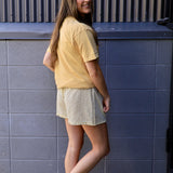 Yellow graphic tee styled with cream plaid shorts and lavender sneakers, worn by smiling brunette model.