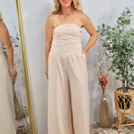 Woman wearing strapless beige and white striped wide-leg jumpsuit with gold jewelry, styled in boho setting with plants and m
