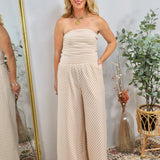 Woman wearing strapless beige and white striped wide-leg jumpsuit with gold jewelry, styled in boho setting with plants and m