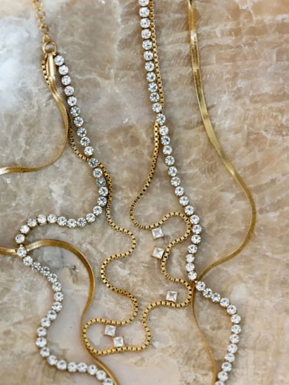 Gold rhinestone tennis necklace, box chain with crystal accents, and herringbone chain layered on marble surface