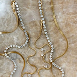 Gold rhinestone tennis necklace, box chain with crystal accents, and herringbone chain layered on marble surface