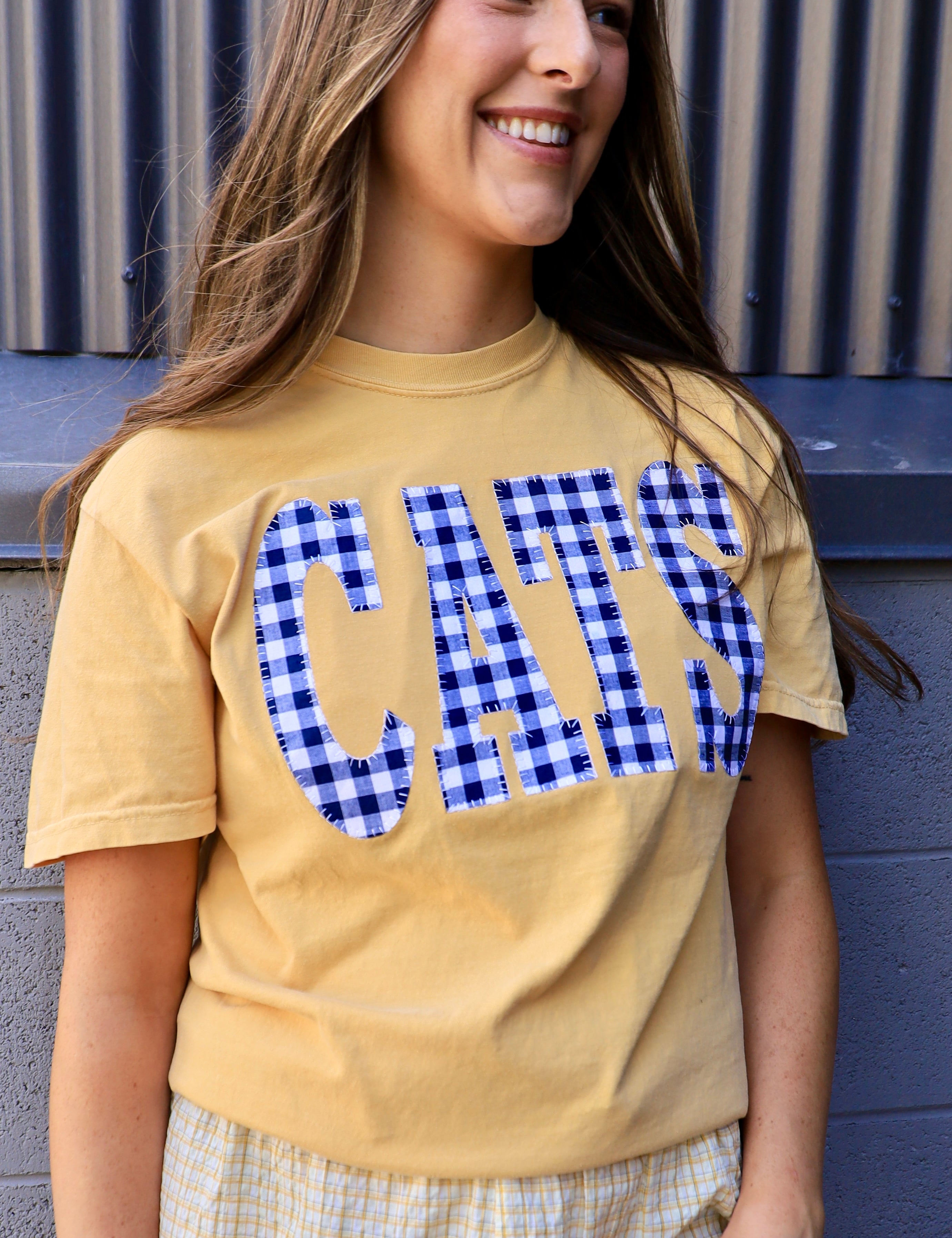 Woman wearing a butter yellow tee with blue gingham CATS applique lettering, smiling outdoors