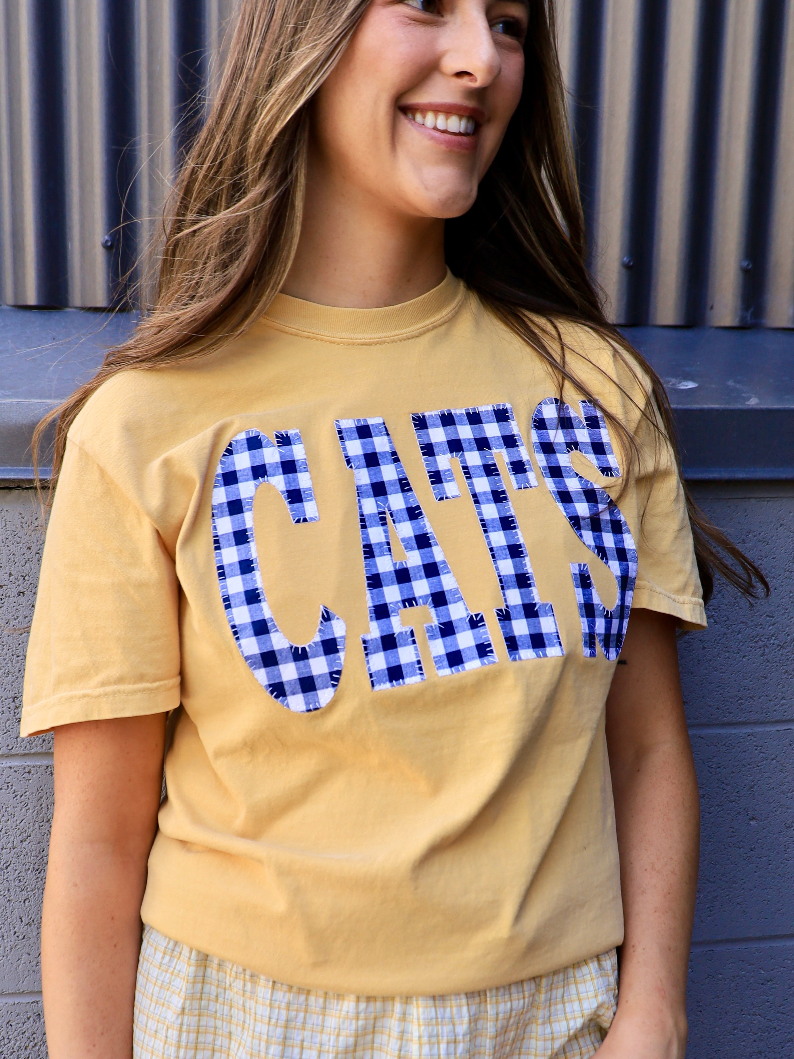 Woman wearing a butter yellow tee with blue gingham CATS applique lettering, smiling outdoors