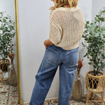Blonde woman wearing cream open-knit sweater with blue jeans, shown from behind in boho-styled room with plants.