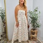 Woman wearing cream strapless midi dress with colorful floral print, styled with white boots and gold jewelry in boho setting