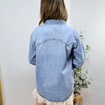 Woman wearing oversized light blue denim shirt from behind, paired with white lace-trimmed shorts in minimalist room setting.