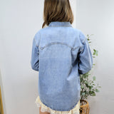 Woman wearing oversized light blue denim shirt from behind, paired with white lace-trimmed shorts in minimalist room setting.