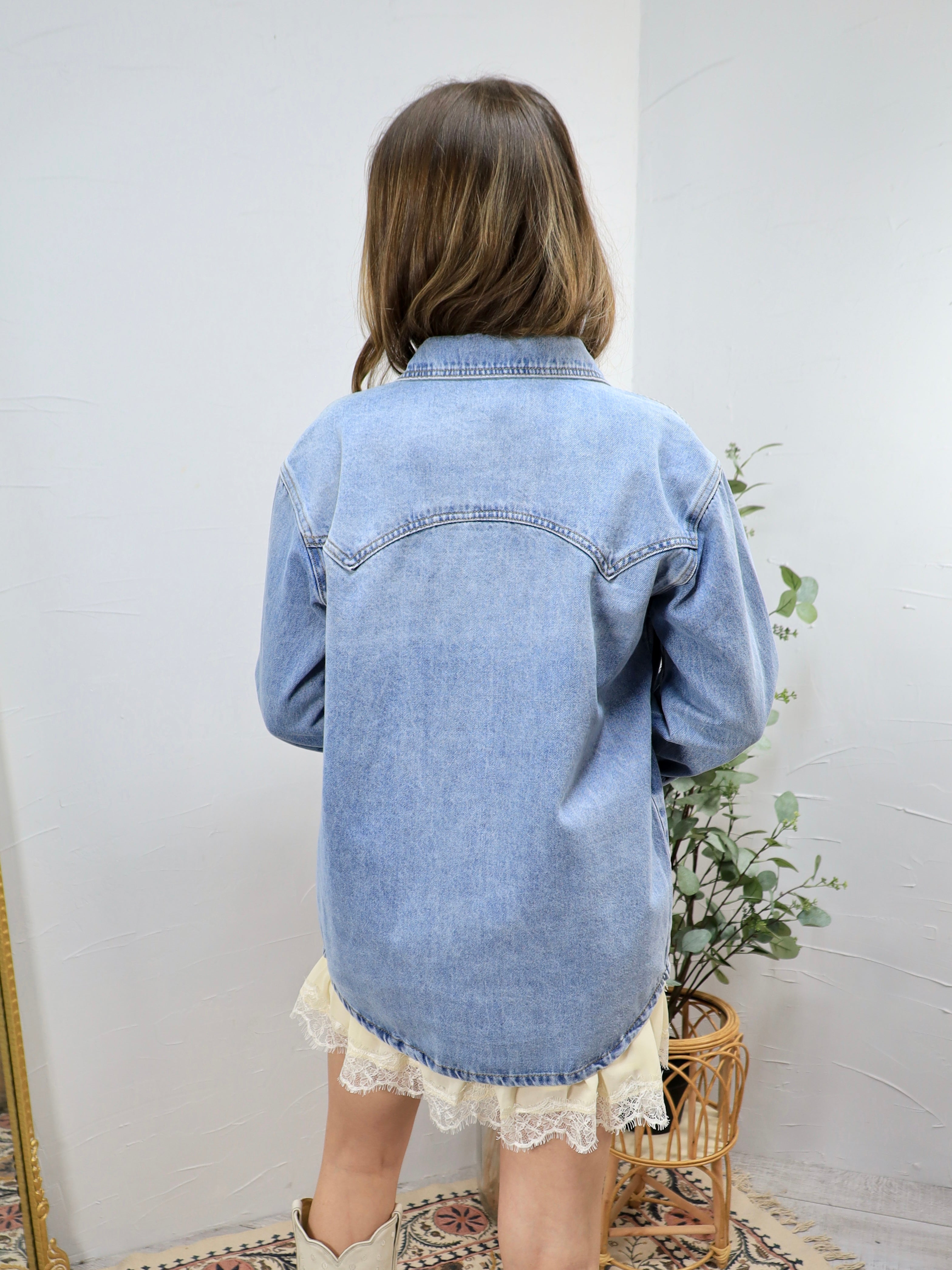 Woman wearing oversized light blue denim shirt from behind, paired with white lace-trimmed shorts in minimalist room setting.
