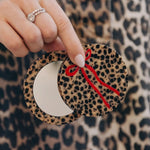 Leopard print compact mirror with red bow detail, held open to reveal mirror, against matching leopard print background.