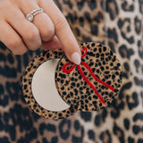 Leopard print compact mirror with red bow detail, held open to reveal mirror, against matching leopard print background.