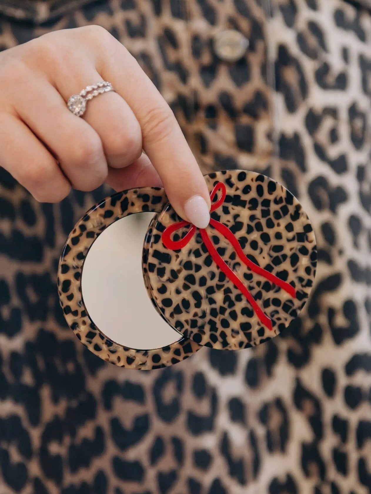Leopard print compact mirror with red bow detail, held open to reveal mirror, against matching leopard print background.
