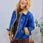 Blue denim jacket with tan sherpa teddy lining and collar, styled open over brown turtleneck and wide-leg pants.