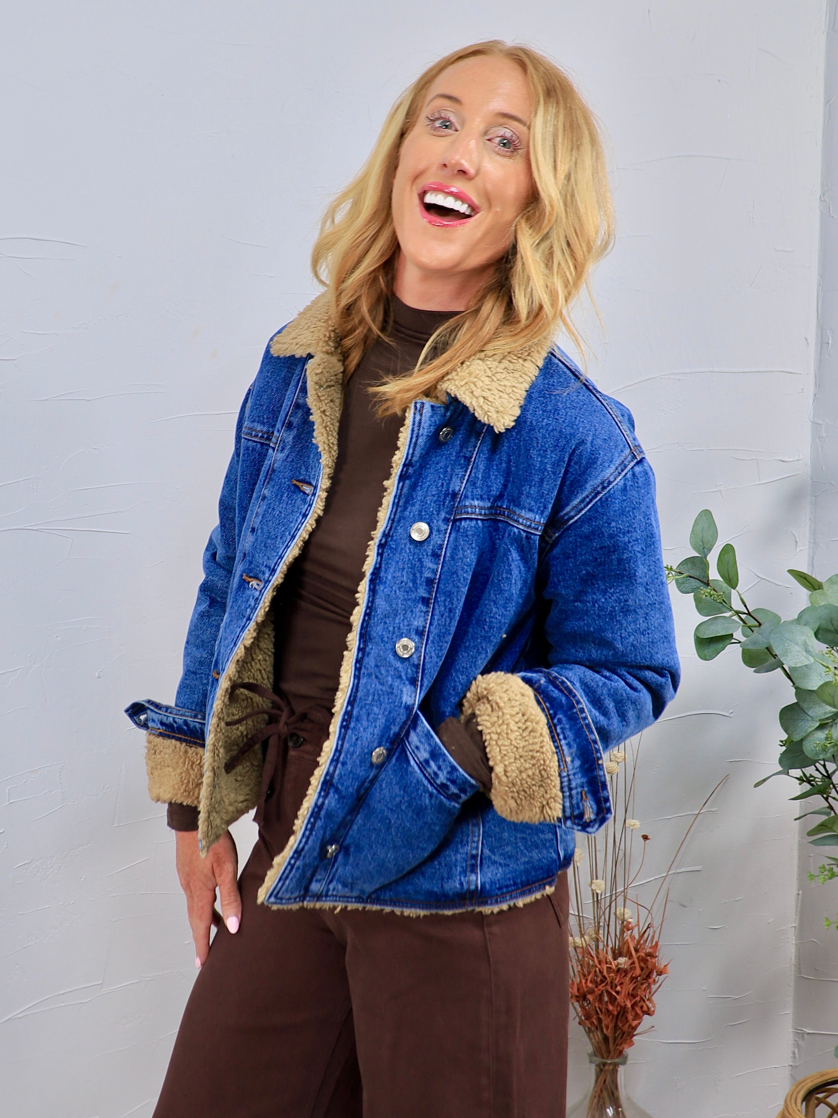 Blue denim jacket with tan sherpa teddy lining and collar, styled open over brown turtleneck and wide-leg pants.