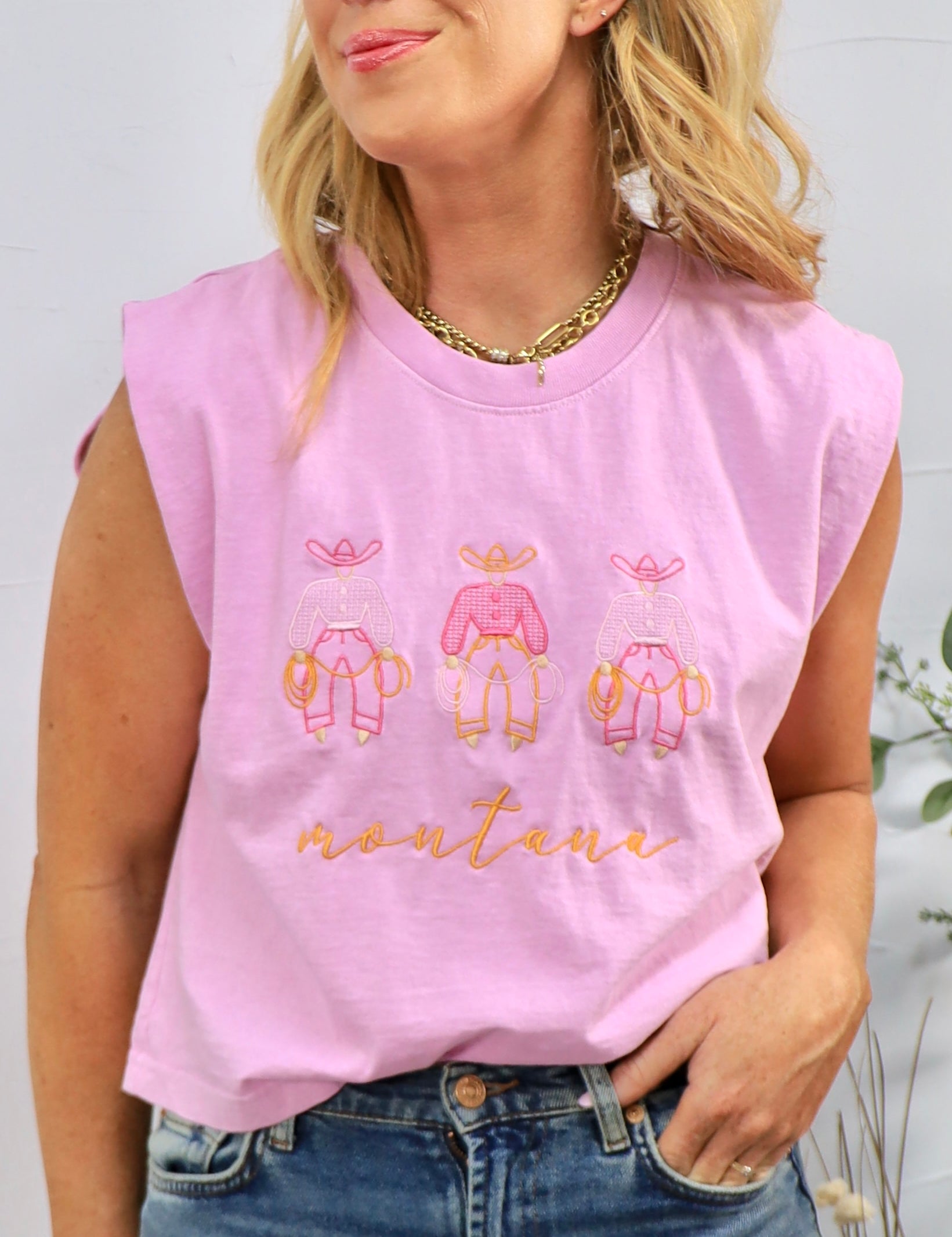 Woman wearing pink sleeveless muscle tee with three cowboy figures and "montana" text, styled with jeans and gold chain neckl