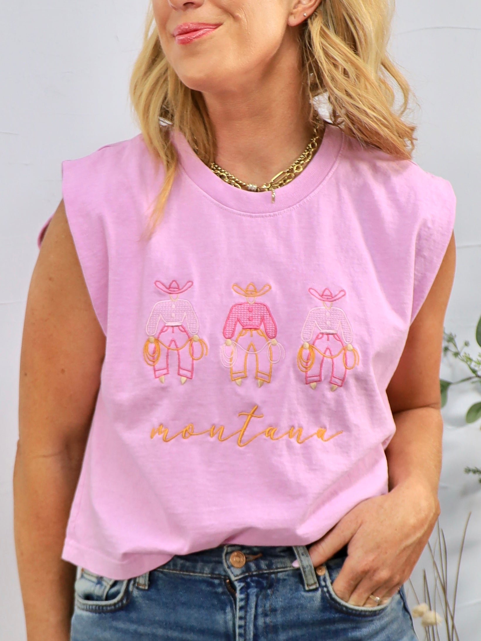 Woman wearing pink sleeveless muscle tee with three cowboy figures and "montana" text, styled with jeans and gold chain neckl