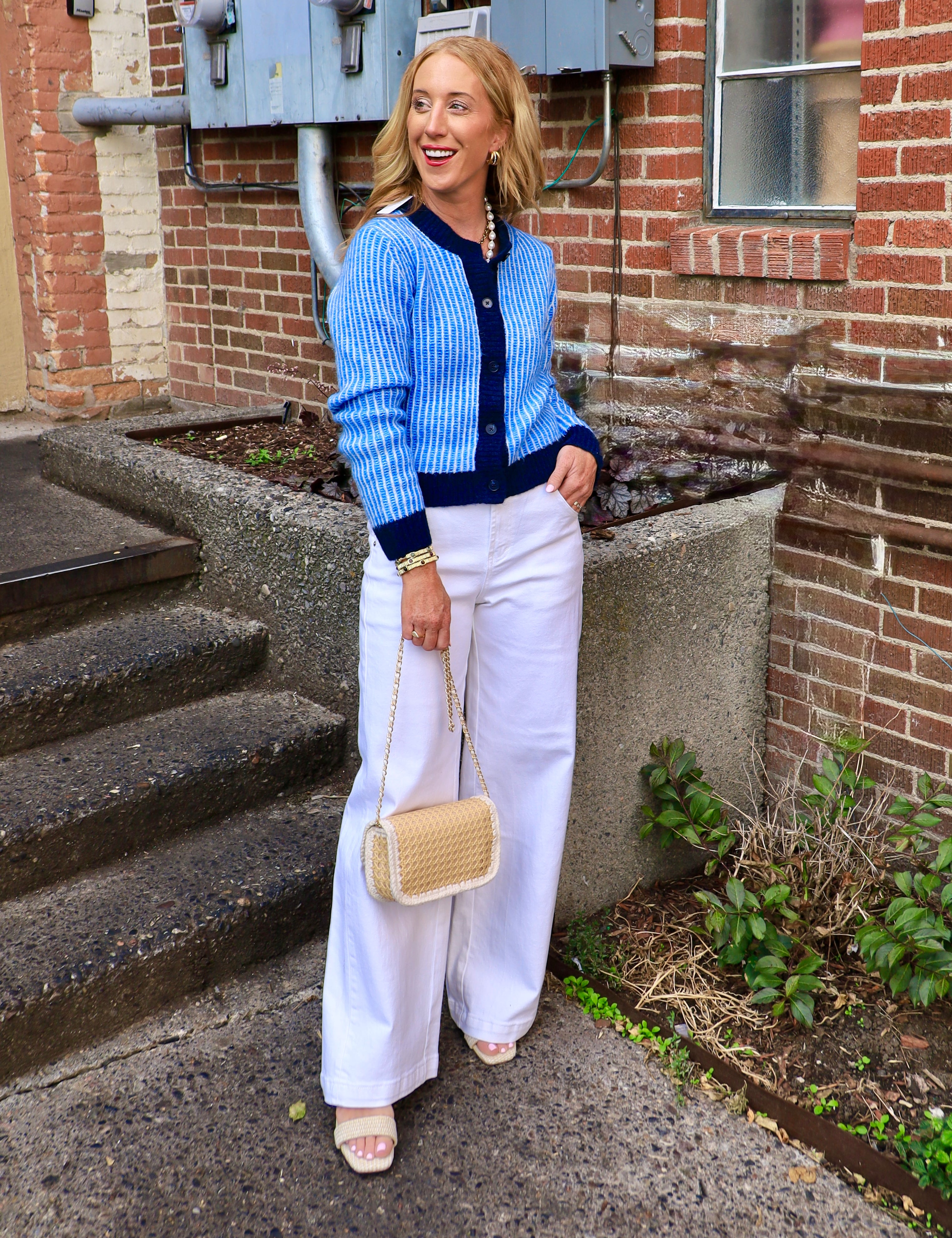 Woman wearing a blue and navy striped fitted button-front cardigan with white wide-leg pants and a woven crossbody bag