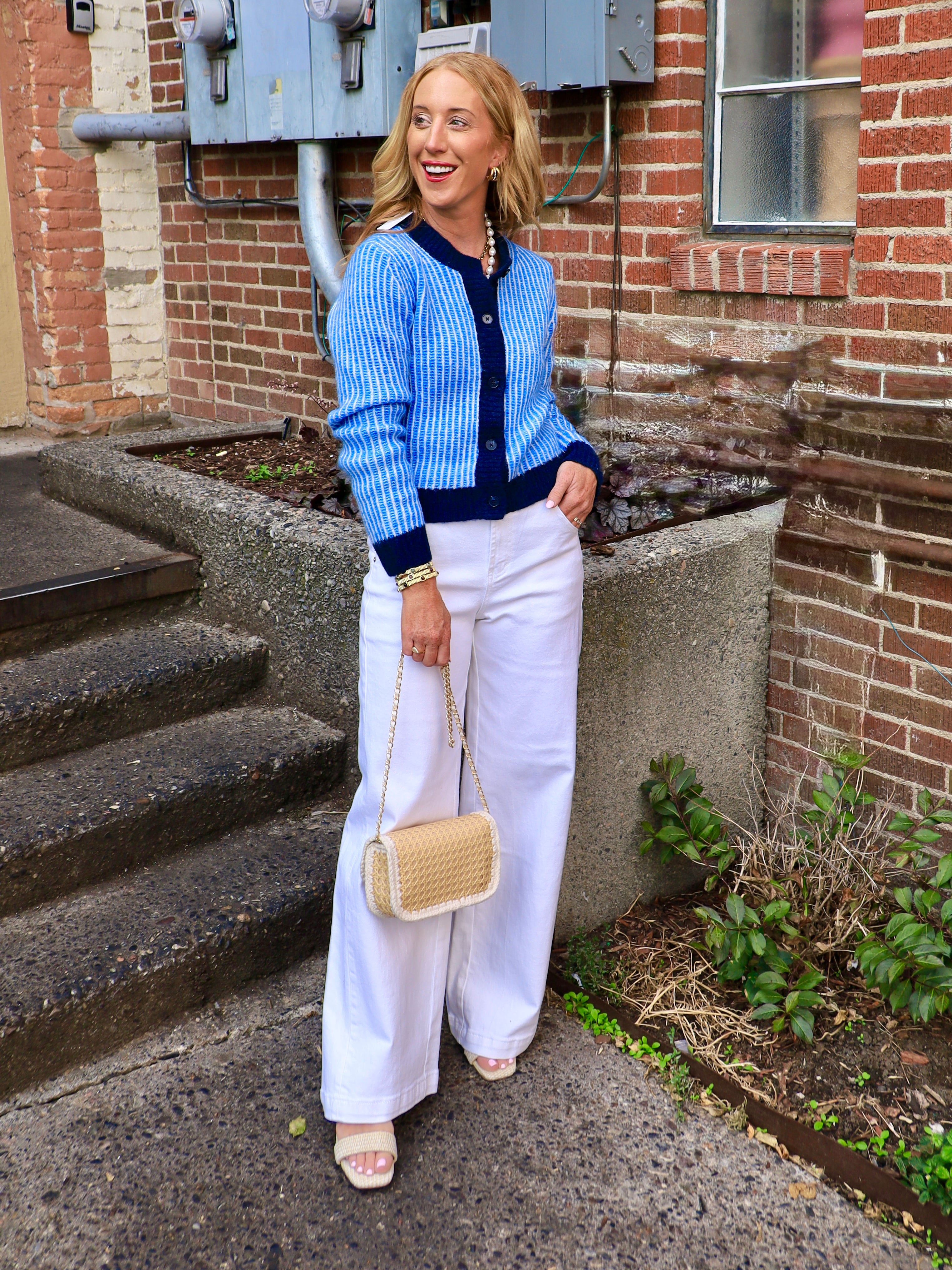 Woman wearing a blue and navy striped fitted button-front cardigan with white wide-leg pants and a woven crossbody bag
