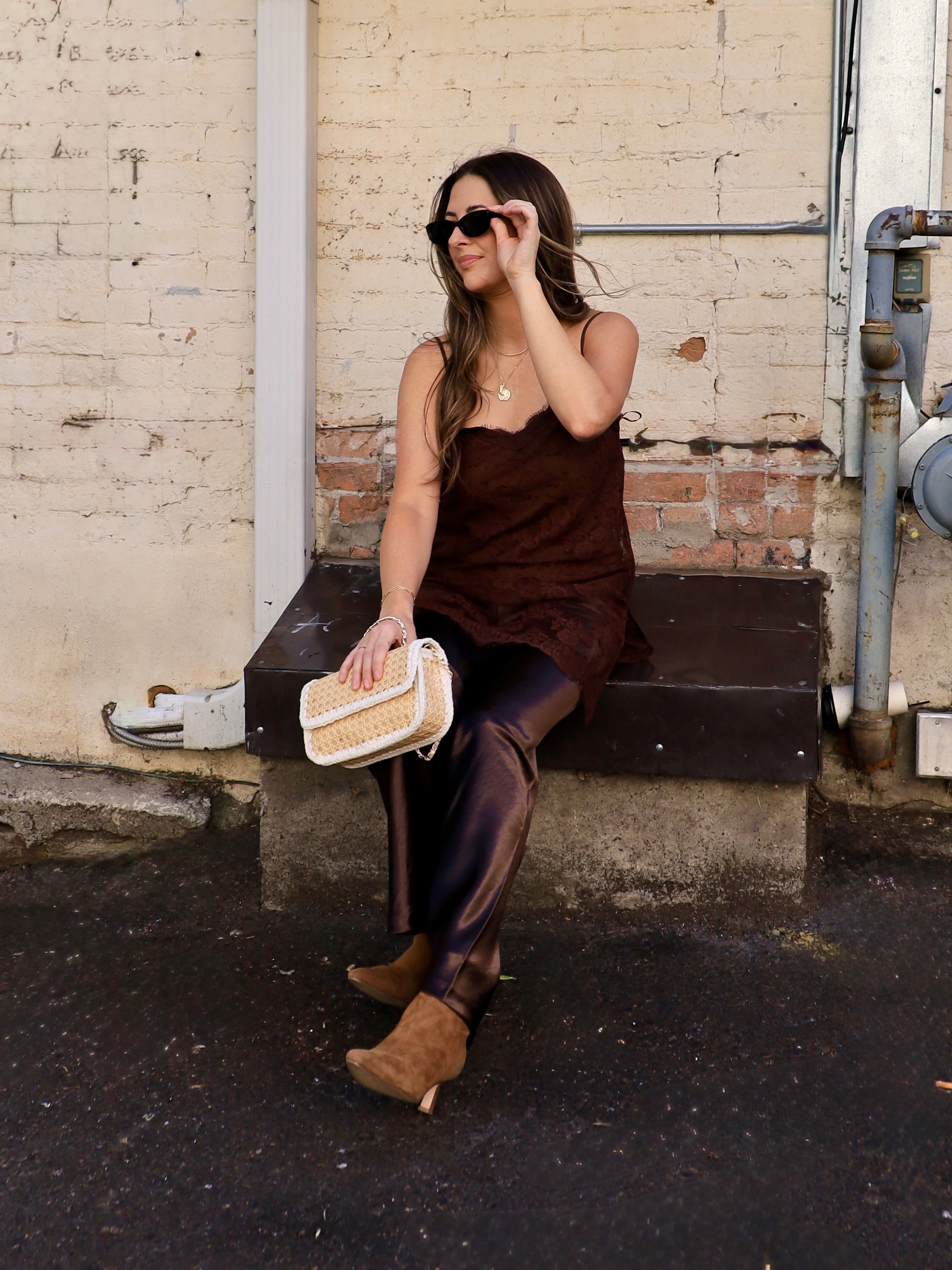 Woman wearing a chocolate brown lace cami with satin trousers and tan suede booties, seated outdoors