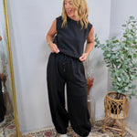 Black sleeveless jumpsuit with drawstring waist and wide-leg pants, styled with silver sneakers.