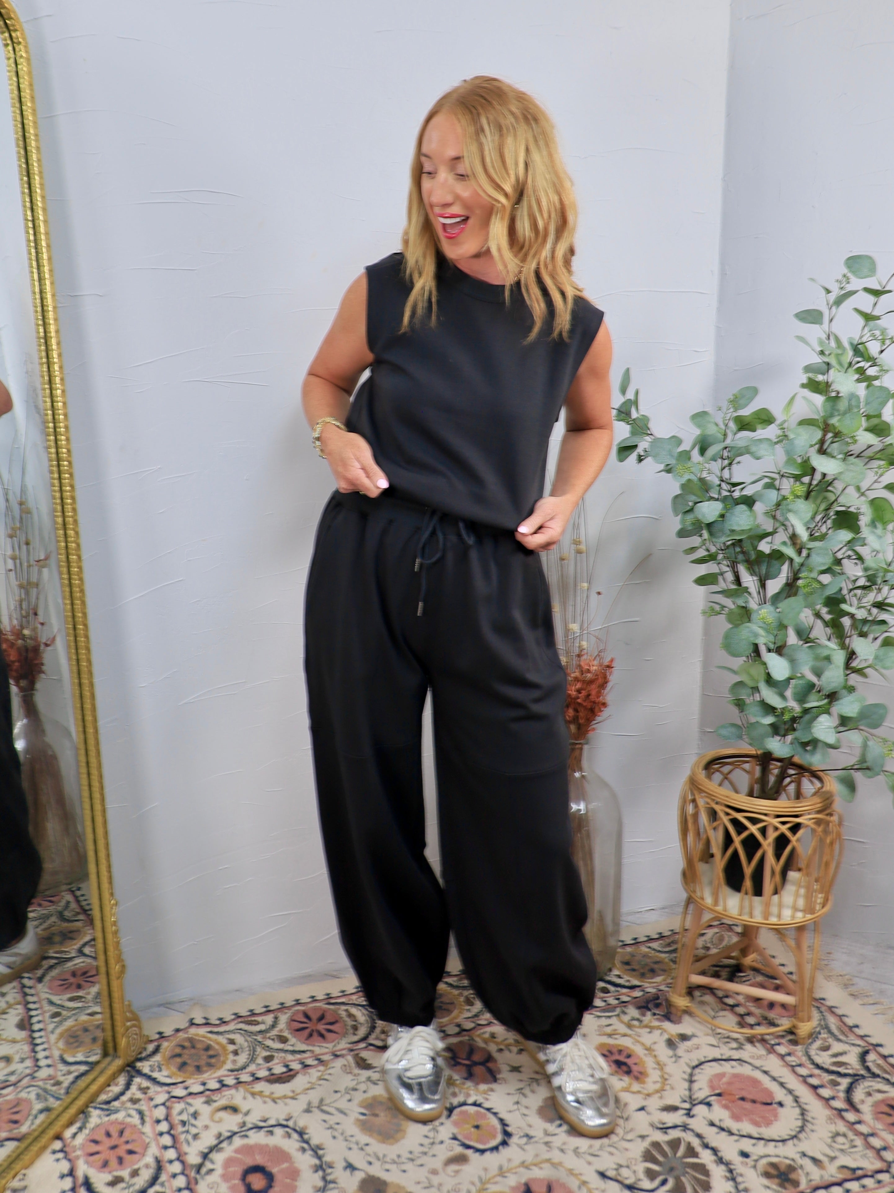 Black sleeveless jumpsuit with drawstring waist and wide-leg pants, styled with silver sneakers.