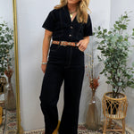 Woman wearing black short-sleeve denim coveralls with brown leather belt and yellow pointed-toe shoes, styled with layered ne